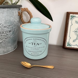 May include: A light blue ceramic tea canister with a lid, featuring the text "TEA" and "The Original Suffolk Canister." A gold-colored spoon rests in front of the canister. The canister is approximately 5 inches tall.