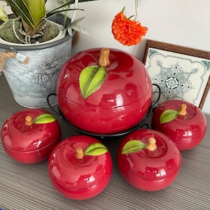 Puede incluir: Conjunto de cinco recipientes de cerámica roja en forma de manzana con hojas verdes. El recipiente más grande está sobre un soporte de metal negro. Los recipientes son perfectos para guardar objetos pequeños como azúcar, té o especias.