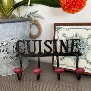 May include: Black metal kitchen hook rack with the word "CUISINE" in large letters. The rack has four hooks with red ceramic knobs.