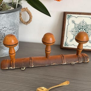 May include: A vintage wooden coat rack with three rounded pegs. The rack is a warm brown color with signs of wear, suggesting age. A decorative tile and a small spoon are also visible.