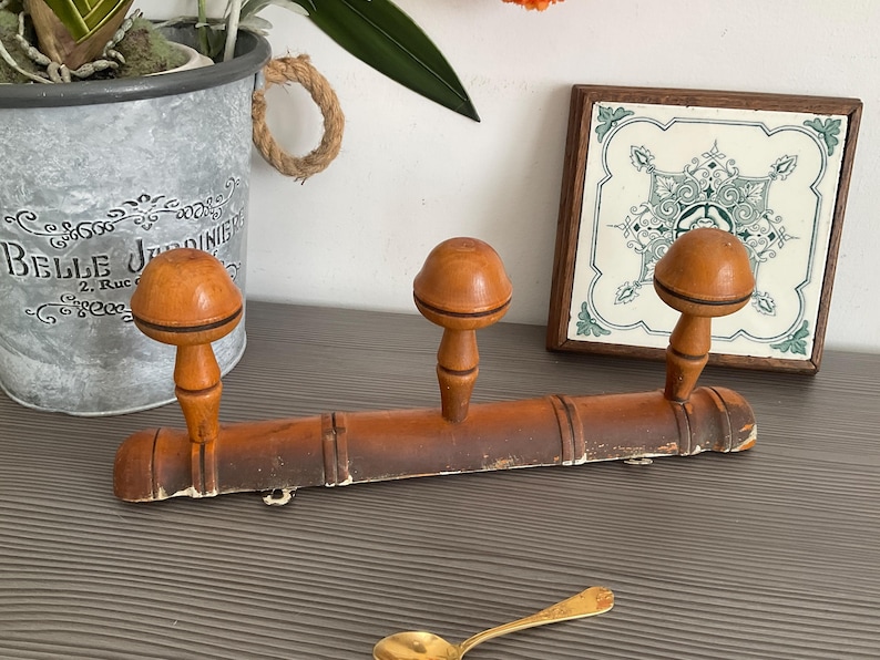 May include: A vintage wooden coat rack with three rounded pegs. The rack is a warm brown color with signs of wear, suggesting age. A decorative tile and a small spoon are also visible.