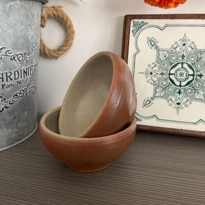 May include: Two small, stacked, brown ceramic bowls with a light beige interior. The bowls have a rounded shape and a slightly textured surface. The image also shows a decorative metal bucket with text and a decorative tile with a floral design.