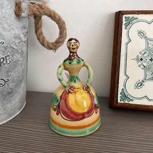 Puede incluir: Una campana de cerámica con forma de mujer que lleva un vestido verde, amarillo y rojo. La campana tiene un diseño floral pintado y una cara sonriente.