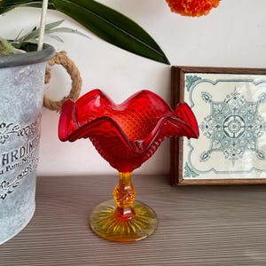 May include: A striking red glass bowl with a wavy rim, supported by a yellow and orange stem and base. The bowl features a diamond pattern. Decorative item.