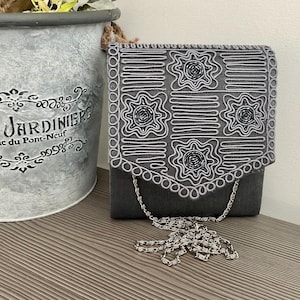 May include: A dark gray rectangular handbag featuring a flap with intricate silver cord embroidery and beaded star-like motifs. The bag includes a silver-toned metal chain strap that rests in front of the main body, which has a smooth texture.