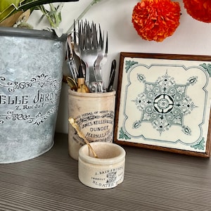 Könnte beinhalten: Eine antike Keramikdose mit einem goldenen Löffel im Inneren. Die Dose trägt ein Etikett mit der Aufschrift "Dundee Marmalade" und ist mit einem grün-weißen Blumendesign verziert. Die Dose steht auf einer Holzoberfläche neben einer Fliese mit einem grün-weißen Blumendesign.