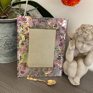Peut inclure: Un cadre photo décoratif avec un motif floral rose, violet et or. Une petite cuillère dorée repose sous le cadre. Une petite statue de chérubin est sur le côté droit du cadre. Un pot en métal avec une plante est à gauche.