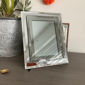 May include: A rectangular picture frame featuring a multi-layered mirrored border with bevelled edges. The frame has a central glass pane surrounded by reflective, angled glass panels that create a stepped, geometric design with a polished finish.