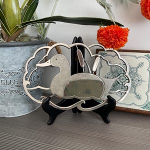 May include: Silver-plated metal hot pad shaped like a duck with a textured surface. The duck is swimming in a decorative frame with a floral design. The hot pad is on a black stand.