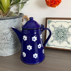 May include: A dark blue enamel coffee pot with a lid and handle. The pot is decorated with white daisy-shaped flowers. It is displayed on a wooden surface, with a decorative tile and a potted plant in the background.