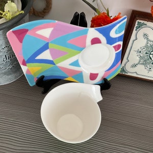 May include: A white ceramic teacup and saucer set with a colorful geometric design. The saucer has a wavy edge and features a variety of colors, including pink, blue, green, and yellow.