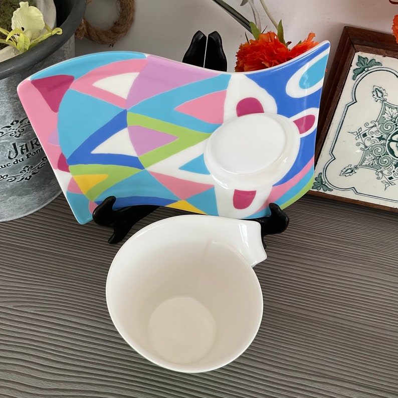 May include: A white ceramic teacup and saucer set with a colorful geometric design. The saucer has a wavy edge and features a variety of colors, including pink, blue, green, and yellow.