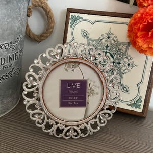 May include: Ornate silver photo frame with a circular aperture. The frame features a decorative, swirling pattern with embedded crystals. The frame holds a photo with the text "LIVE FRAME" and dimensions of 9cm x 9cm.
