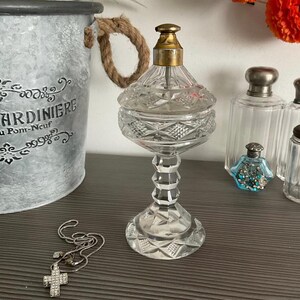 Pode incluir: Uma garrafa de vidro transparente com um padrão de diamante e uma tampa dourada. A garrafa está sobre uma superfície cinza com outras garrafas de vidro.