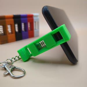 Könnte beinhalten: Ein grüner, 3D-gedruckter Telefonständer mit Federmechanismus, der ein schwarzes Smartphone hält. Der Ständer ist an einem Schlüsselanhänger mit einem silbernen Verschluss befestigt. Weitere farbige Ständer sind sichtbar.
