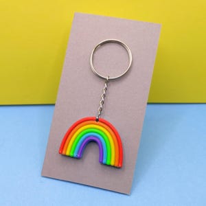 May include: A rainbow-shaped keyring with a silver ring. The rainbow design features red, orange, yellow, green, blue, and purple arches. The keyring is attached to a silver chain and is displayed on a grey background.