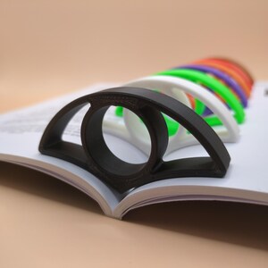 May include: A black plastic page holder shaped like an eye, with a ring in the centre. The page holder is on top of a book, with other page holders in different colours visible behind it.