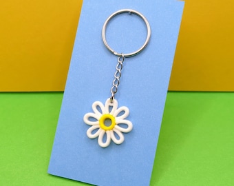 Peut inclure: Un porte-clés en forme de marguerite avec une fleur blanche et un centre jaune, attaché à une chaîne et un anneau argentés. Le porte-clés est présenté sur une carte bleue sur un fond jaune et vert.