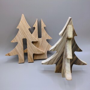 May include: Wooden Christmas tree decorations in various sizes. The trees are made of light-coloured wood with visible grain patterns. The trees are cut into the shape of a fir tree, with a flat base for standing.