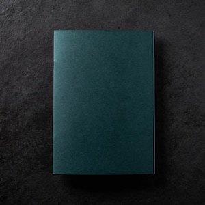 Könnte beinhalten: Ein teal-farbenes Notizbuch mit glatter, matter Oberfläche. Das rechteckige Notizbuch ist auf einer dunklen, strukturierten Oberfläche zentriert. Der Einband hat eine durchgehende, tiefe Teal-Farbe.