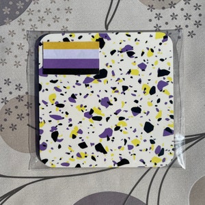 May include: A white coaster with a yellow, purple and black terrazzo pattern. A small yellow, white and purple flag is adhered to the coaster.