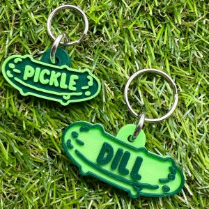 May include: Two green pickle-shaped keyrings, one with the word "PICKLE" and the other with "DILL" in dark green lettering. Each keyring has a silver metal ring attached for keys.