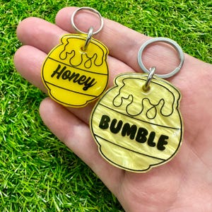 Może przedstawiać: Dwa żółte breloki w kształcie słoika miodu z czarnym tekstem. Jeden brelok ma napis "Honey", a drugi "Bumble".
