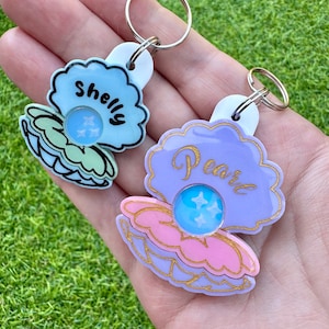 May include: Two keychains shaped like seashells. The top keychain is light blue with the word "Shelly" written on it. The bottom keychain is purple with the word "Pearl" written on it.