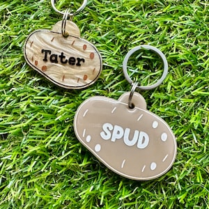 May include: Two brown potato-shaped dog tags with silver rings. The top tag says "Tater" and the bottom tag says "Spud".