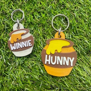 Könnte beinhalten: Zwei Schlüsselanhänger mit Honigtöpfen mit den weißen Wörtern "WINNIE" und "HUNNY". Die Honigtöpfe sind braun mit gelbem Honig, der von oben tropft. Die Schlüsselanhänger liegen auf einer grünen Grasfläche.