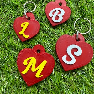May include: Four red heart-shaped keyrings, each with a different initial letter: L, B, M, and S. The letters are yellow or white. Each keyring has a silver ring attached. The keyrings are displayed on green grass.