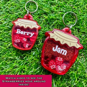 May include: Two red glass jar-shaped keyrings with glitter and small red strawberries inside. The jars have gold lids and say "Berry" and "Jam" on them.