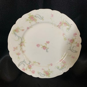 May include: A white porcelain plate with a delicate floral pattern in shades of pink and blue. The plate has a scalloped edge and a raised rim.