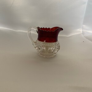 May include: A clear glass creamer with a red glass rim and a scalloped edge. The creamer has a textured design and a small handle.