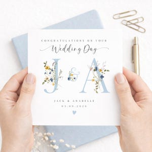 May include: A wedding card held by hands, with the words "Congratulations on your Wedding Day." The card shows the initials "J & A" in pale blue, decorated with floral details. The names "Jack & Anabelle" and the date "03.09.2026" are also displayed.