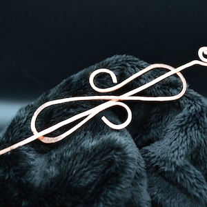 May include: A copper hair stick with a decorative swirl design. The stick is thin and has a long, slender handle.