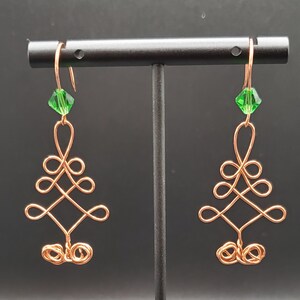 May include: A pair of copper wire earrings with green crystal accents. The earrings are shaped like a stylized tree with a diamond-shaped center.