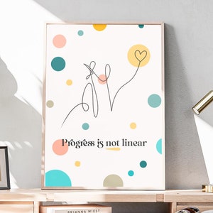 May include: A white framed poster with colorful dots and a line drawing of a heart with the text "Progress is not linear". The poster is hanging on a white wall above a wooden desk with a lamp, a plant, and books.