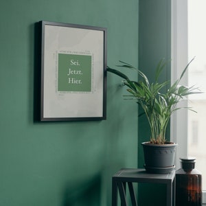 K&ouml;nnte beinhalten: Ein gerahmter Druck mit dem Text "Sei. Jetzt. Hier." in einem gr&uuml;nen Farbschema. Der Druck h&auml;ngt an einer gr&uuml;nen Wand neben einer Topfpflanze und einem kleinen Tisch.
