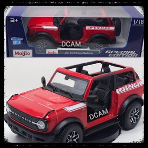 May include: A red 1/18 scale die-cast metal model of a 2021 Ford Bronco Badlands. The model has black wheels, a white stripe, and the word "LIFEGUARD" on the side. The model is in its original box with the "Maisto" logo.