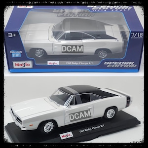 May include: A white 1969 Dodge Charger R/T die-cast metal model car with a black roof and racing stripes. The model is 1/18 scale and features the text "DCAM" on the side. The model is displayed on a black base and in a box.
