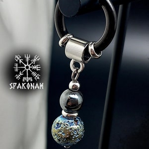 Peut inclure: Une boucle d'oreille argentée avec un charme en pierre noire et bleue. Le charme est attaché à un anneau argenté avec une perle argentée. La boucle d'oreille est sur un support noir.