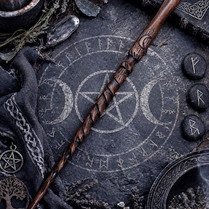 May include: A dark brown wooden wand featuring a spiral carved handle and a crescent moon symbol near the top. The wand is positioned diagonally across a circular stone slab engraved with a pentagram, moon phases, and runic characters.