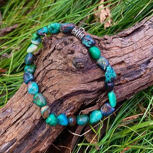 May include: A turquoise and green gemstone bracelet with a silver clasp. The bracelet is made of smooth, rounded stones and is laid out on a piece of wood.