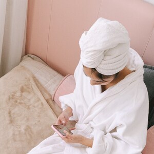 May include: A woman in a white bathrobe sits on a bed with a pink laptop, a camera, and photos. She is using a smartphone.