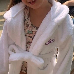 May include: A white hooded robe with a purple embroidered "y/a" on the left side. The robe is tied at the waist with a white sash. The person wearing the robe is smiling and has a black and white checkered headband.