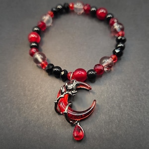May include: A beaded bracelet featuring alternating black, clear, and red spherical beads. A crescent moon pendant with a red translucent centre and a dark metal frame hangs from the bracelet, accented by a small red teardrop-shaped charm.