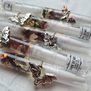 May include: A set of clear glass tubes filled with dried flowers, glitter, and gold leaf. Each tube has a silver bat charm attached to it. The tubes are labelled with the text "DARK DESIRES" and "CITRUS OIL PUMPKIN SPICE".