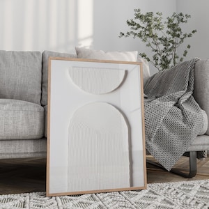 May include: A white framed wall art piece with a raised, textured design of overlapping circles and arches. The art is hanging on a white wall above a gray couch with a gray throw blanket.