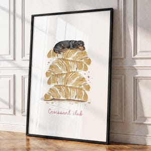 Puede incluir: Una l&aacute;mina de arte enmarcada con una ilustraci&oacute;n en acuarela de un perro salchicha negro y fuego descansando sobre una pila de cuatro cruasanes dorados. El texto "Croissant club" aparece en cursiva rosa bajo la pila de pasteles.